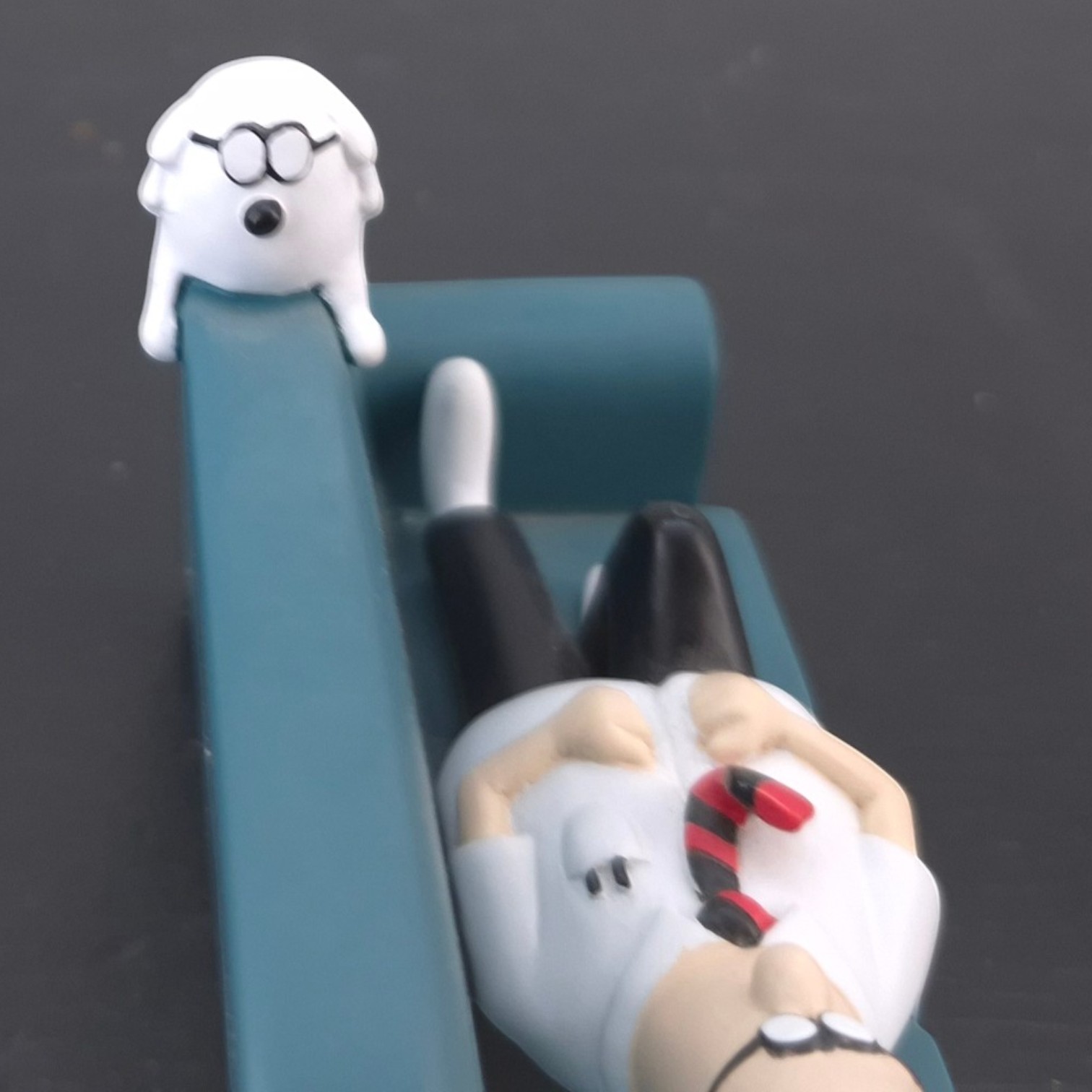 Dilbert and Dogbert Figurine _top - Small white cartoon figure with round glasses sitting on a blue chair, looking surprised.