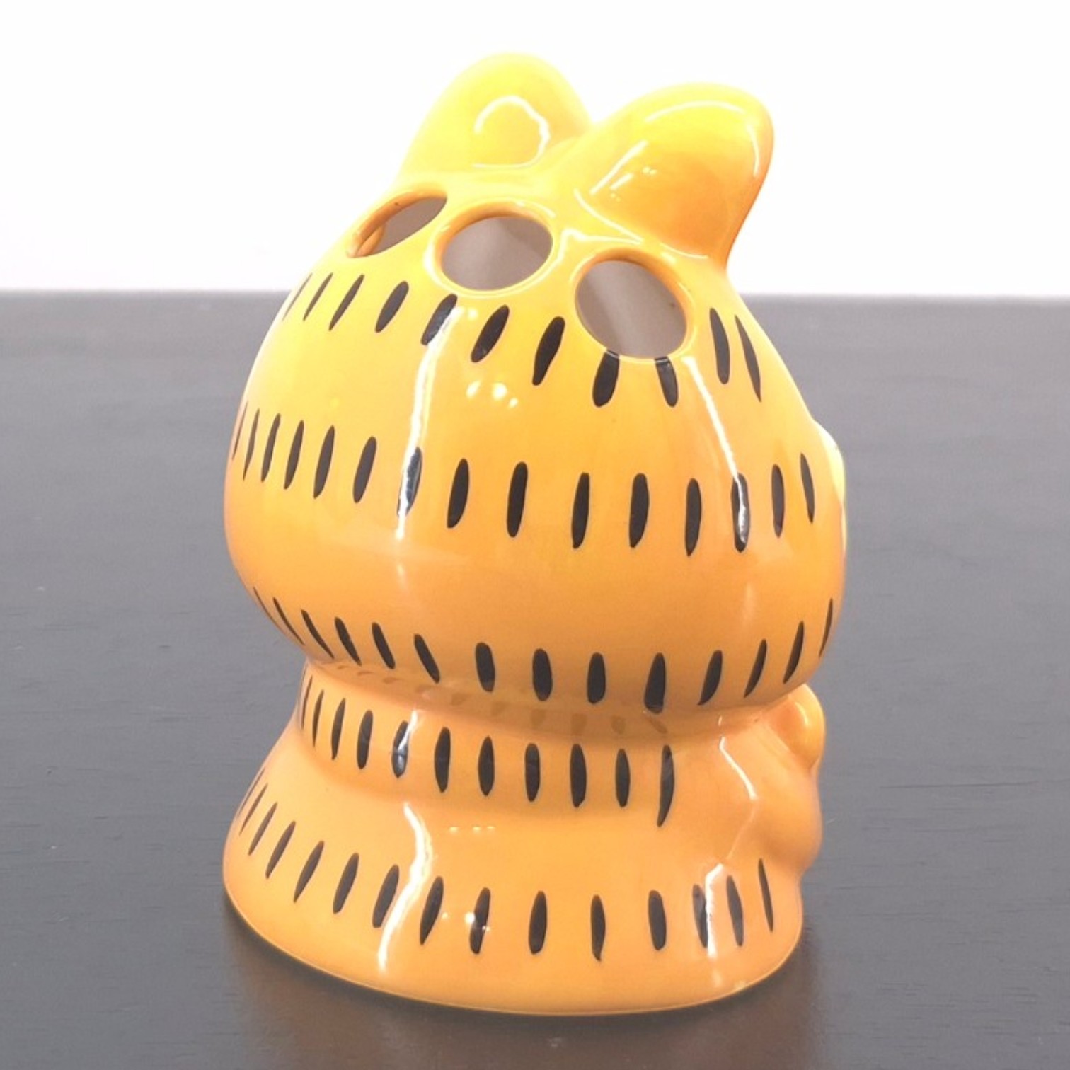 Garfield Toothbrush Holder _right back - Yellow glossy ceramic owl-shaped piggy bank with black vertical stripes and two large eye holes sitting on a dark surface