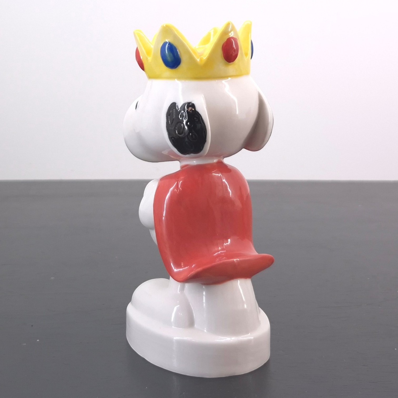King Snoopy Fève _left back - White dog figurine wearing a yellow crown with colorful gems and a red shirt.