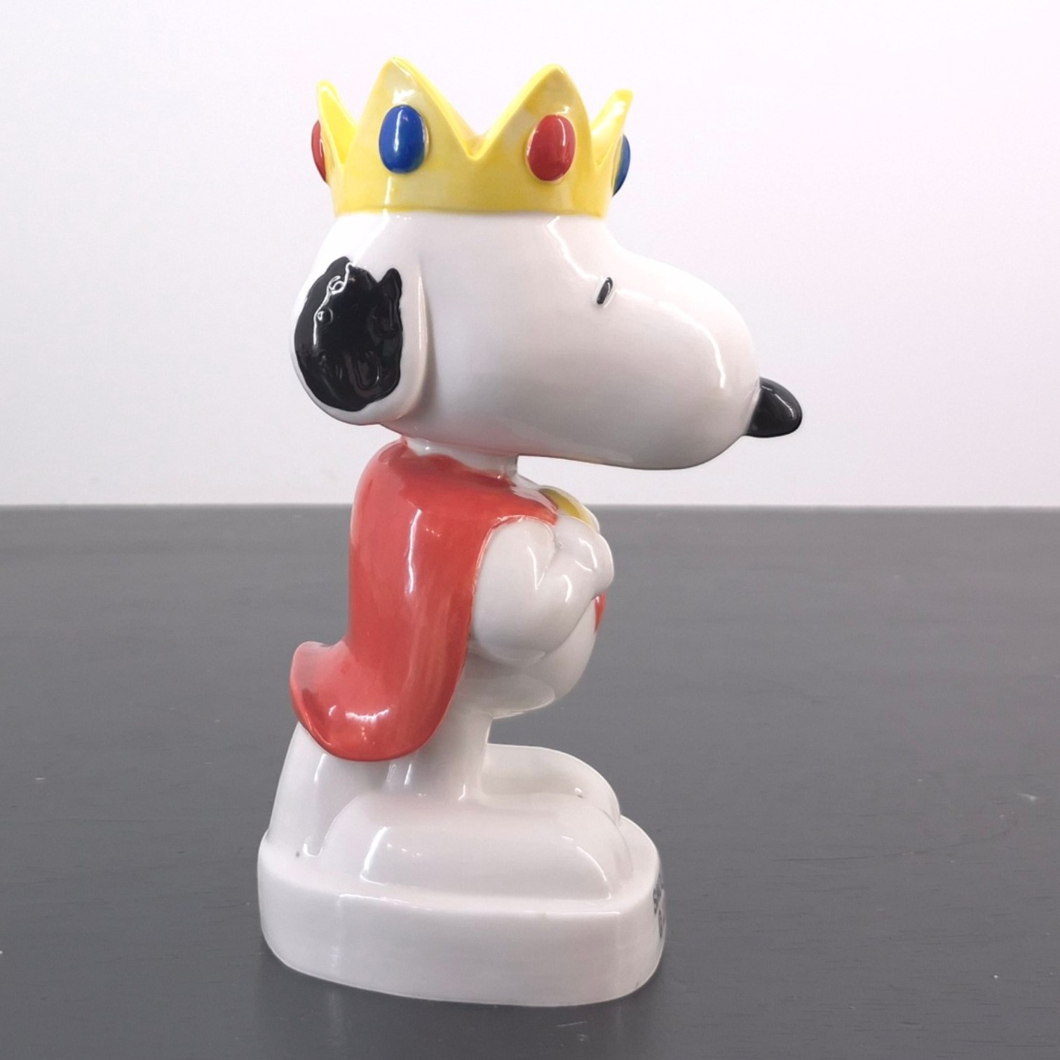 King Snoopy Fève _right - White Snoopy figurine wearing a yellow crown with red and blue gems, standing on a white pedestal against a gray surface.
